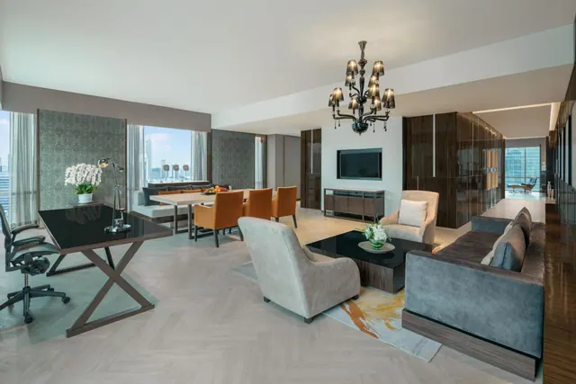 A suite living area with a dining table and orange chairs, a grey sofa, armchairs, a coffee table, a wall-mounted television, a black desk, and large windows showing city buildings outside.