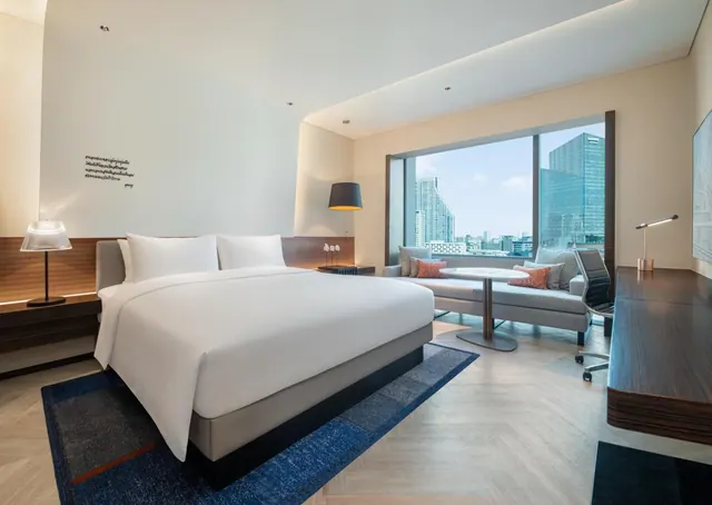 A bedroom with a king bed, a seating area by floor-to-ceiling windows overlooking city high-rises, and a wooden desk on the right.
