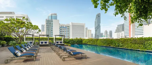 A rooftop swimming pool with blue loungers on a timber deck, with city towers and the twisted Mahanakhon tower in the background.