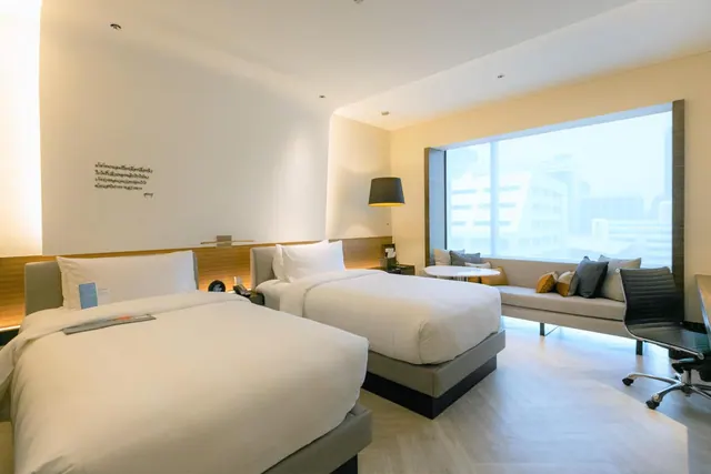 A hotel bedroom with two single beds, a grey sofa and a small round table by a large window looking over city buildings, and a desk with a black chair on the right.