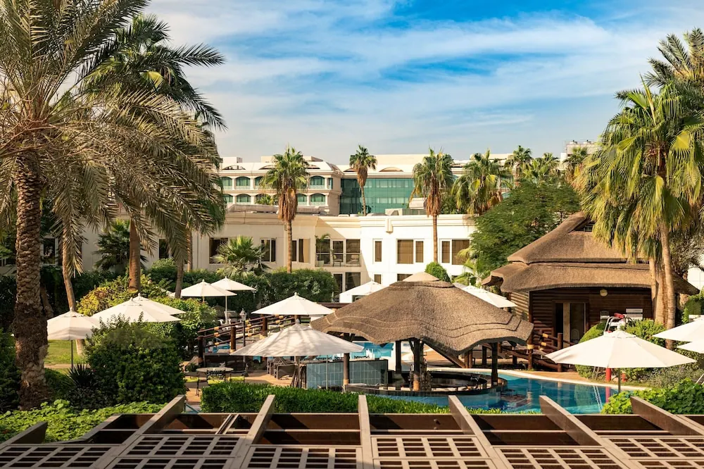 A pool with a thatched-roof pavilion, white parasols and palm trees, seen from a raised terrace, with a white hotel building behind.
