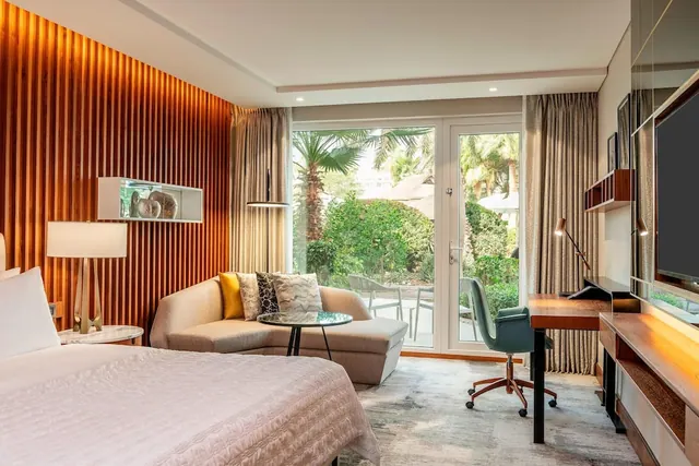 A hotel bedroom with a large bed, a beige sofa with cushions, a desk with a green chair and a wall-mounted television, and glass doors opening onto a garden with palm trees.