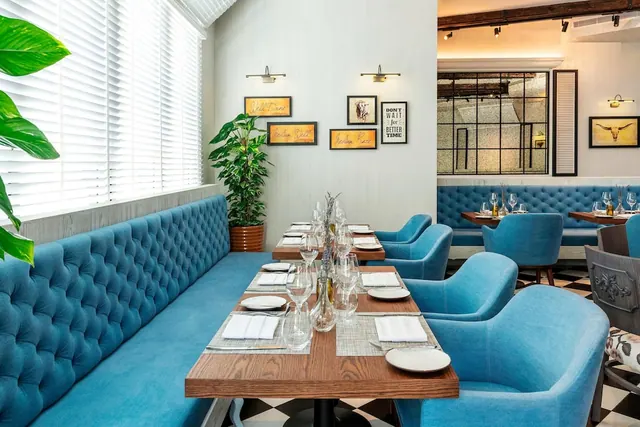 A set wooden dining table with white napkins, plates and glasses sits beside blue tufted banquette seating in a restaurant with a checkered floor.