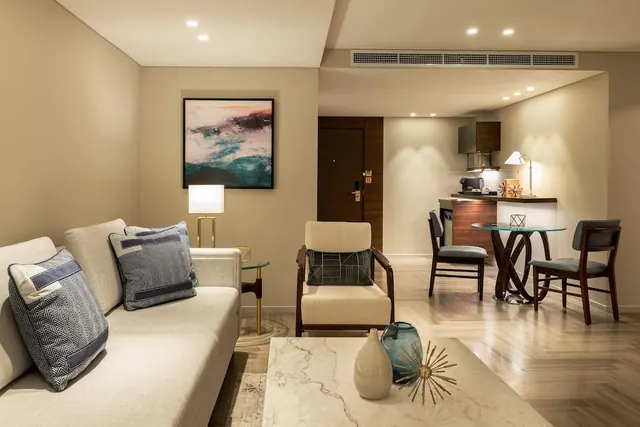 A living area in a hotel suite with a cream sofa, an armchair, a round glass dining table and a small kitchenette with a coffee machine.