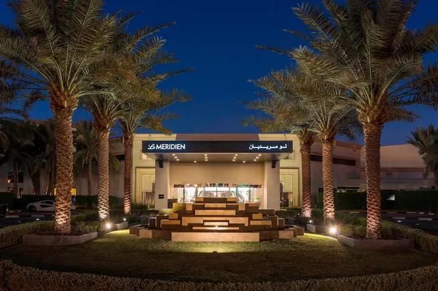 The illuminated entrance of a Le Meridien hotel at dusk, with rows of palm trees and a stepped stone feature in front of the building.