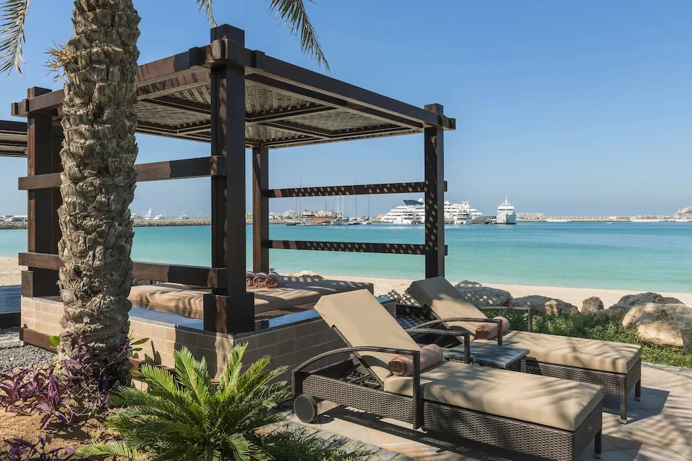 A wooden pergola cabana and two wicker sun loungers sit on a paved terrace beside a sandy beach, with turquoise sea and moored yachts beyond.