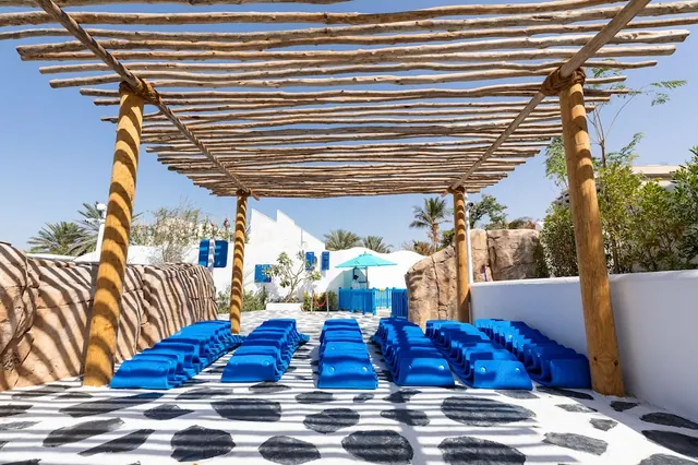 A shaded outdoor area under a wooden pergola with rows of blue loungers on a black-and-white patterned floor, with white walls and a blue umbrella in the background.