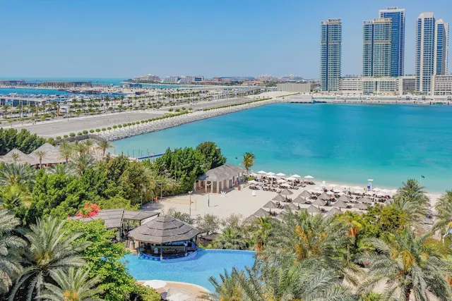 Aerial view of a sandy beach with umbrellas and loungers, a pool with a thatched bar, the turquoise sea and high-rise towers beyond.