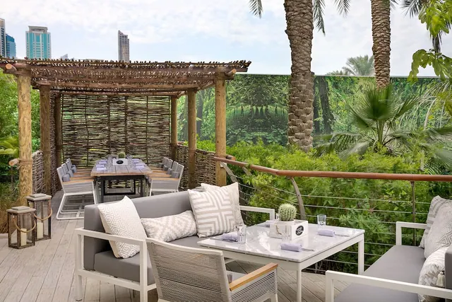 An outdoor terrace with grey sofas, a marble coffee table and a covered dining table under a timber pergola, with palm trees, lush greenery and high-rise towers in the background.
