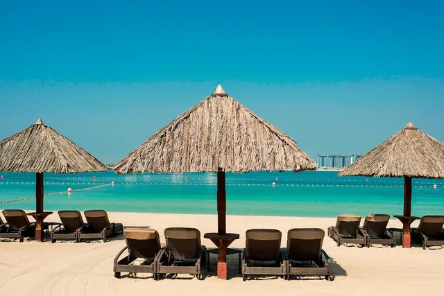 Thatched umbrellas and cushioned sun loungers sit on a sandy beach facing turquoise water marked by a line of buoys.