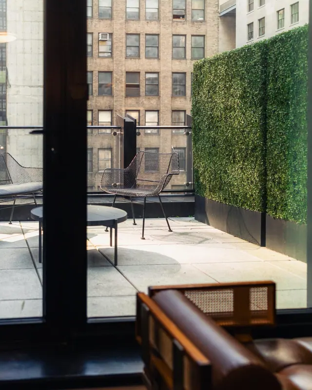 A view through a glass door from an interior room onto a terrace with wire chairs, a round table and a green hedge wall, with brick buildings beyond.