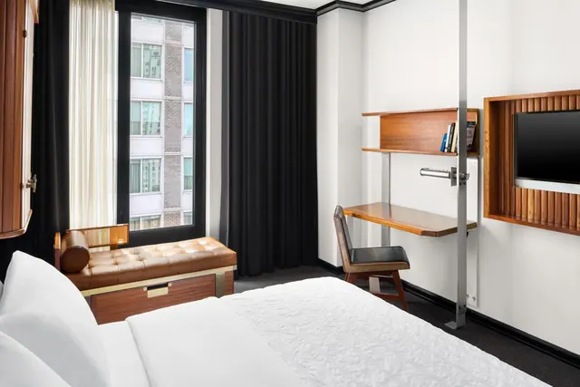 A bedroom with a made white bed, a tufted leather bench by a tall window, a wall-mounted desk with a chair, and a wall-mounted television.