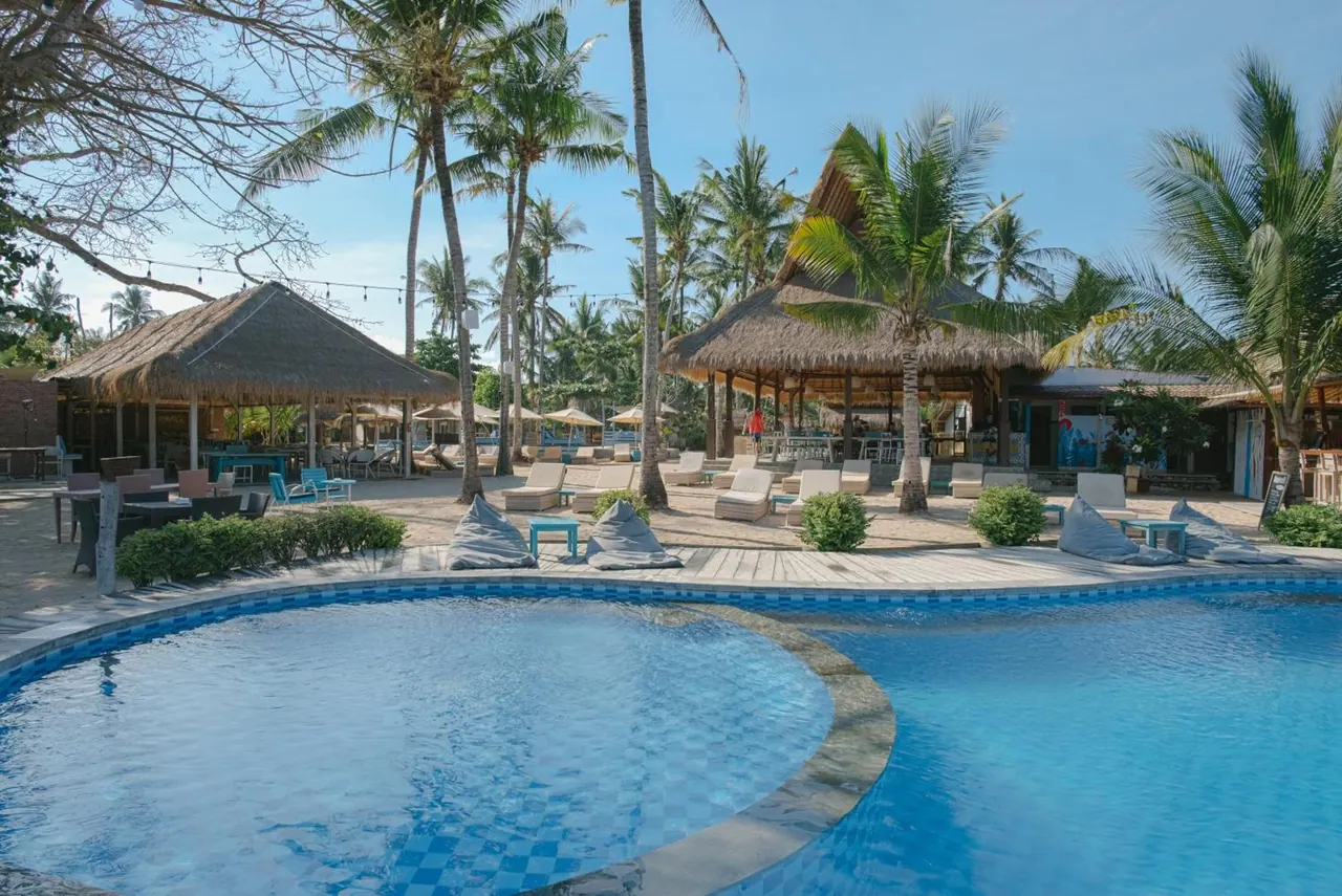 A large curved blue-tiled swimming pool sits in front of thatched-roof bars, sun loungers and palm trees.