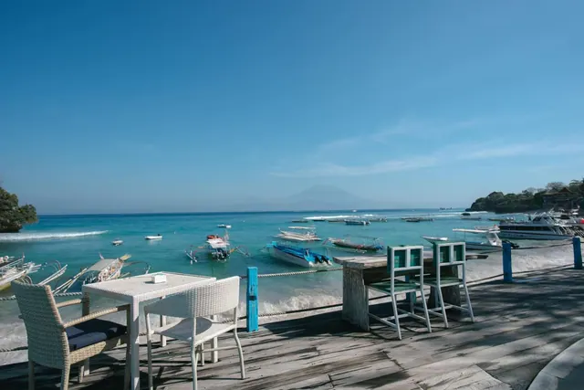 White wicker tables and chairs sit on a weathered wooden terrace overlooking turquoise sea, with traditional outrigger boats moored in the bay and a volcano on the hazy horizon.