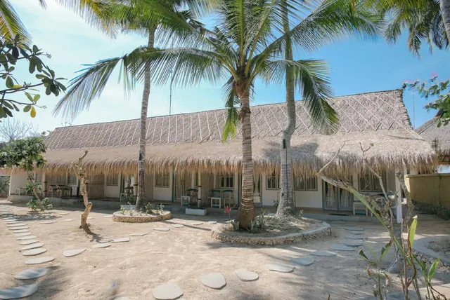A single-storey thatched-roof building with a covered veranda stands behind palm trees and a sandy courtyard with stepping stones.