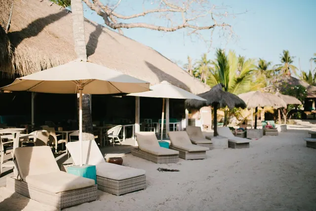 Wicker sun loungers and cream parasols stand on white sand beside thatched-roof buildings and palm trees.