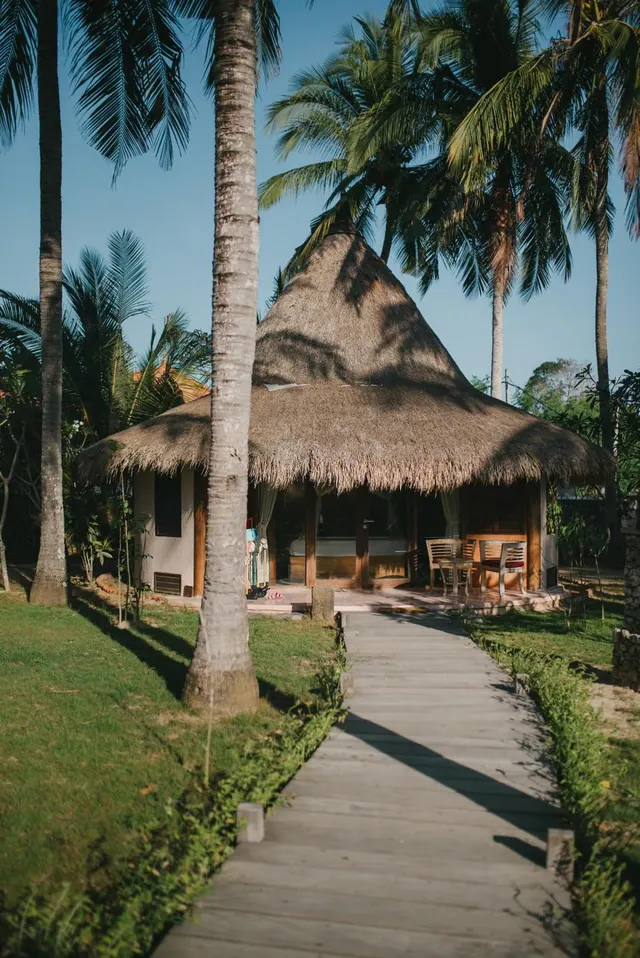A thatched-roof bungalow stands behind a wooden walkway through a lawn, with tall palm trees around it and chairs on its deck.