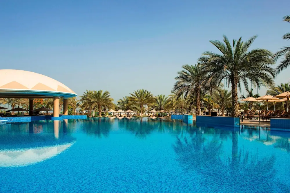 A large blue swimming pool with a domed pool bar on the left, and palm trees and sun loungers under umbrellas on the right.