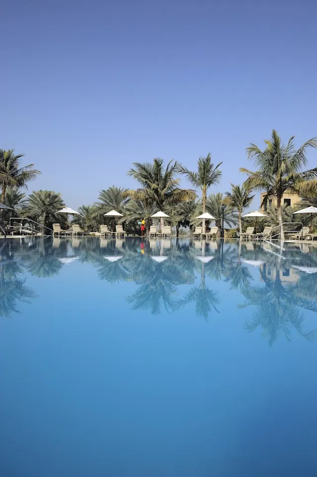 A large outdoor pool with white shade umbrellas, sun loungers and palm trees along the far edge, reflected in the still water.