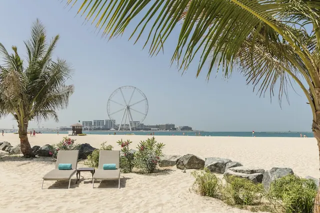 Two grey sun loungers with teal cushions stand on a sandy beach, framed by a palm tree, with the Ain Dubai ferris wheel and a city skyline visible across the water.