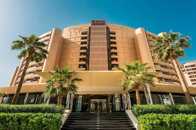 Front exterior of the Royal Meridien hotel building with palm trees, hedges and a flight of steps leading up to the entrance under a clear blue sky.