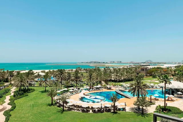 Elevated view over a hotel pool area with rows of loungers and white umbrellas, palm-lined lawns, a sandy beach and the sea beyond.