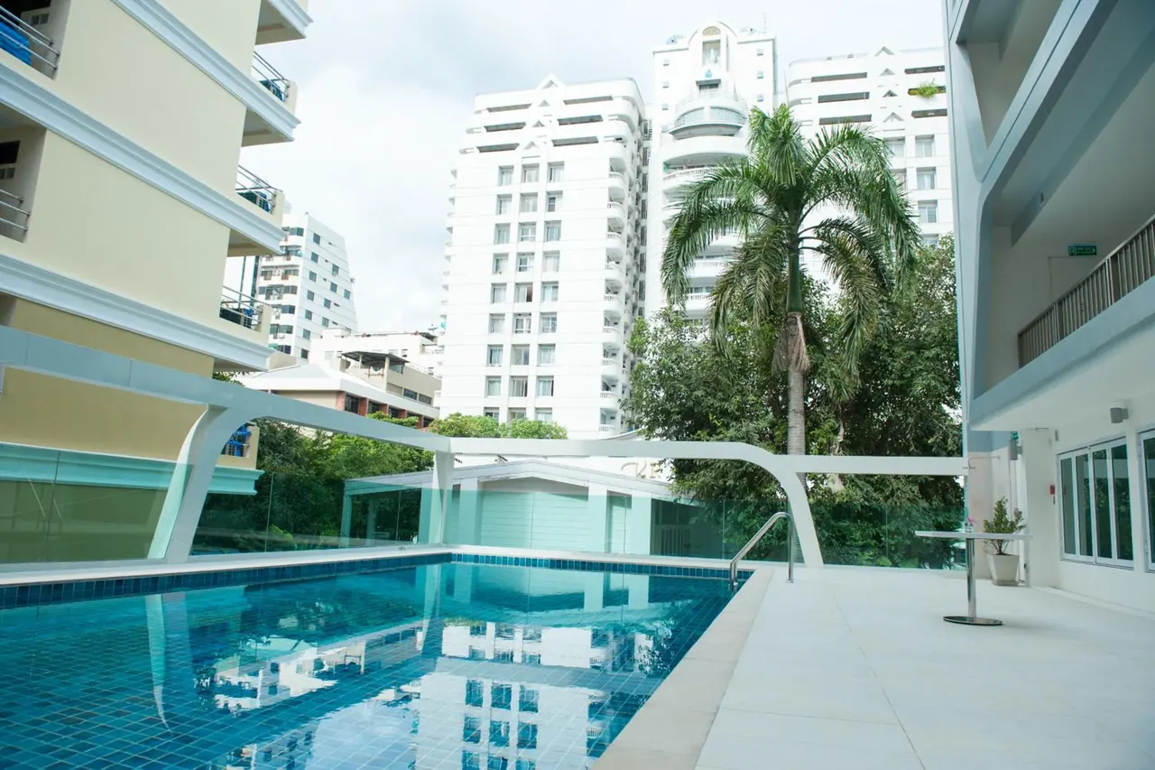 A swimming pool with blue tiled edges on a white deck, with a palm tree and high-rise residential buildings visible beyond the glass railing.