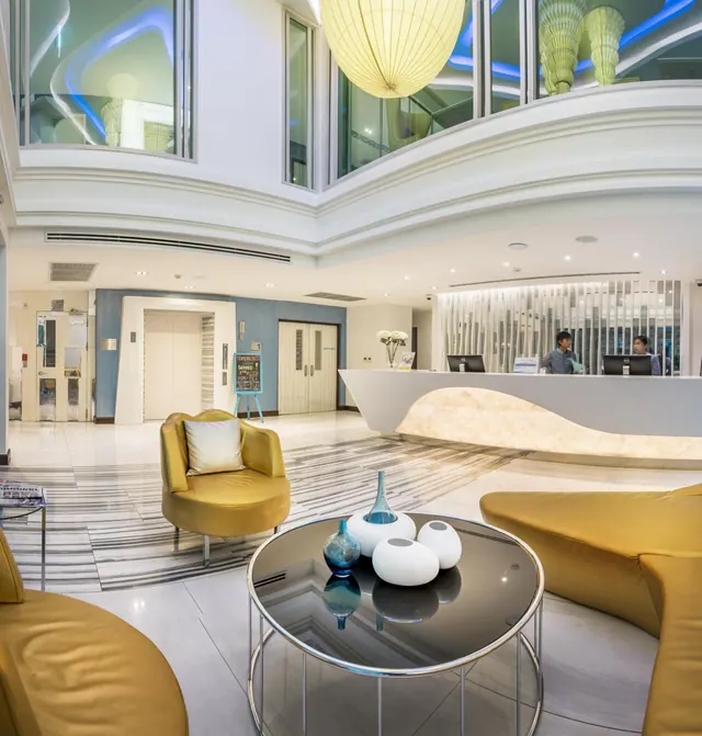 A lobby with yellow armchairs, a glass coffee table with white and blue vases, and a curved reception desk where two staff are working.