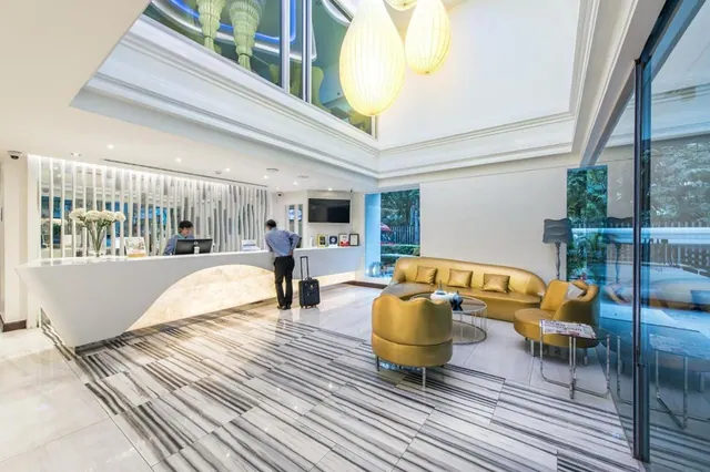 A lobby with a curved white reception desk, a staff member behind it, a guest pulling a suitcase, and gold sofas and armchairs on a striped stone floor, with trees visible through the glass wall.