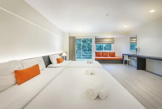 A bedroom with two beds dressed in white linen and orange cushions, an orange window seat, a desk, and a glass door looking out onto trees.