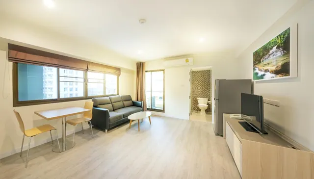 A bright living area with a grey sofa, a round coffee table, a small dining table with two chairs, a television on a wooden cabinet and a fridge, with the bathroom visible through an open doorway.
