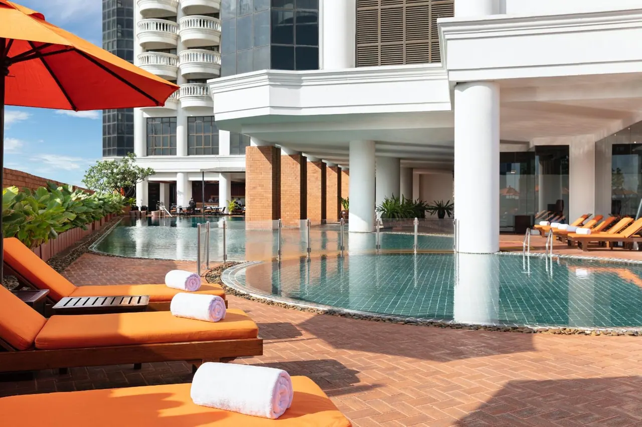 Poolside area with orange sun loungers, rolled white towels and an orange umbrella beside a tiled pool under white columns.