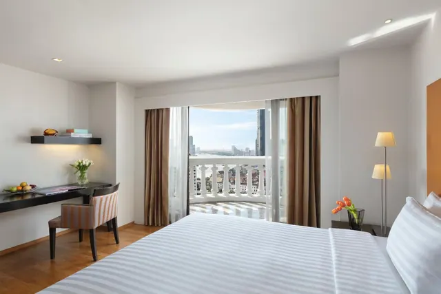 A bedroom with a made bed, a desk and chair, and sliding glass doors opening onto a balcony with a view of a river and city buildings.