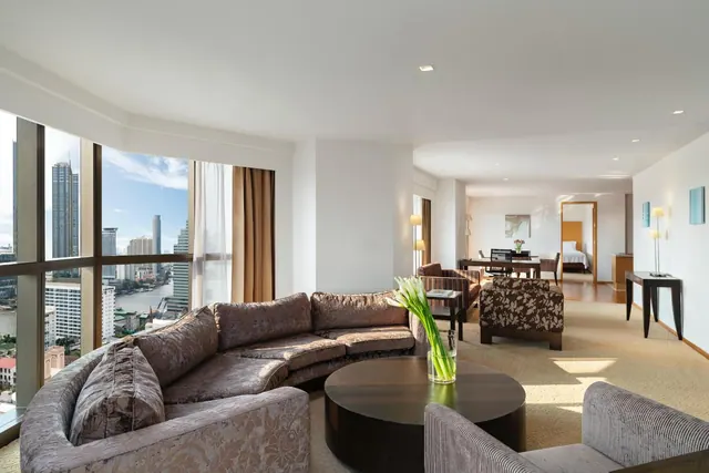 A suite living room with a curved sofa, a round coffee table holding a vase of white flowers, and floor-to-ceiling windows overlooking a river and city high-rises.