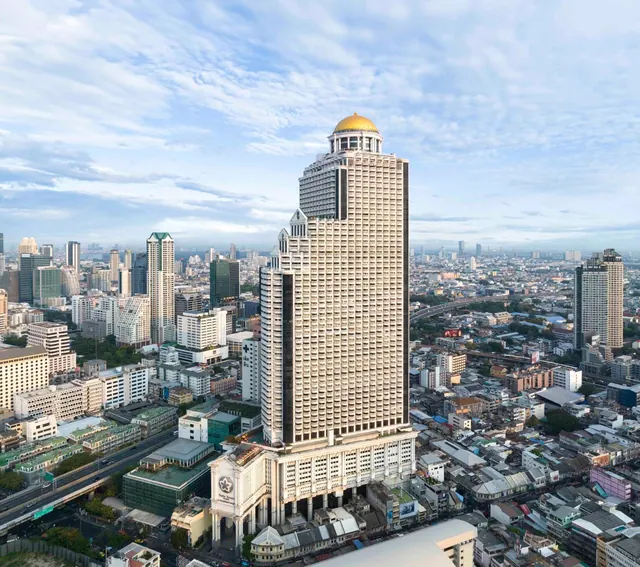 Aerial view of a tall tower with a golden dome rising above a dense city of mid-rise buildings under a partly cloudy sky.