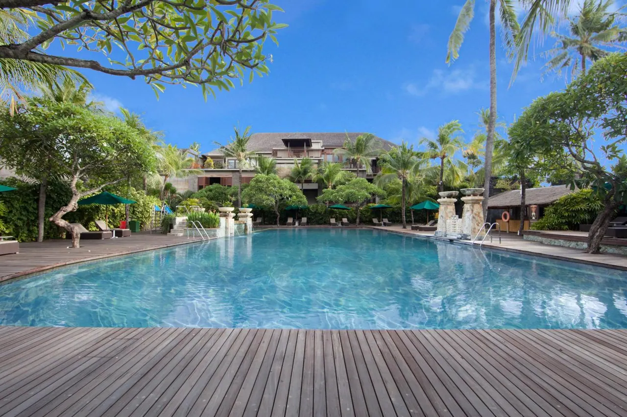A large outdoor swimming pool with a wooden deck, sun loungers and green umbrellas, backed by a multi-storey hotel building and palm trees.