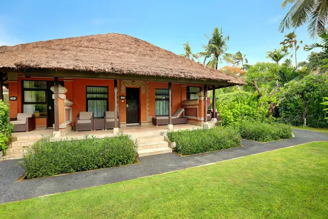 A single-storey Balinese-style villa with a thatched roof and orange walls, with a veranda holding wicker chairs, set among a lawn, palm trees and tropical plants.