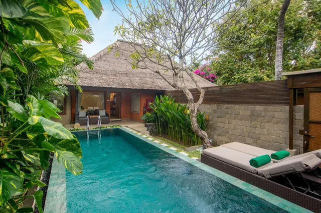 A narrow private swimming pool sits beside a thatched-roof villa, with two sun loungers holding green towels on the right.