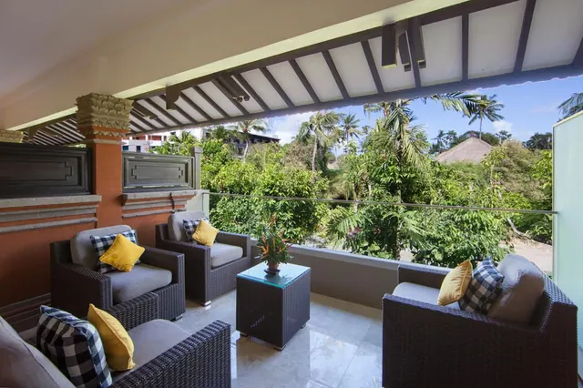 A covered balcony with wicker armchairs, yellow and checked cushions, a glass side table with a potted plant, and a view over tropical trees and palms.
