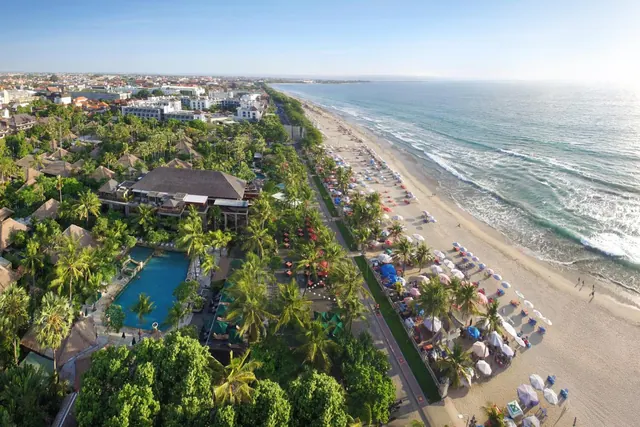 Aerial view of a beachfront resort with a swimming pool, tropical gardens, rows of beach umbrellas and loungers on the sand, and the ocean beyond.