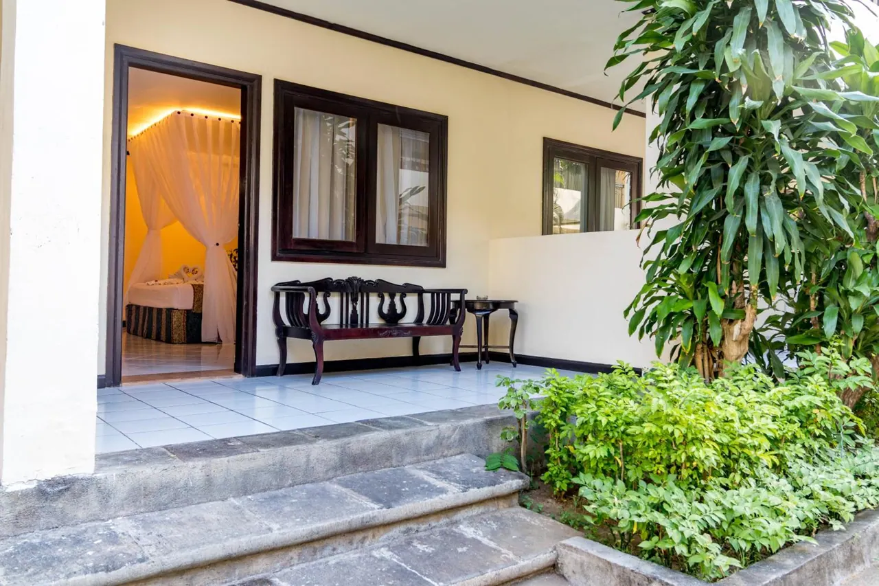 A carved wooden bench sits on a tiled veranda beside an open doorway into a bedroom with a canopy bed, with leafy plants at the right.