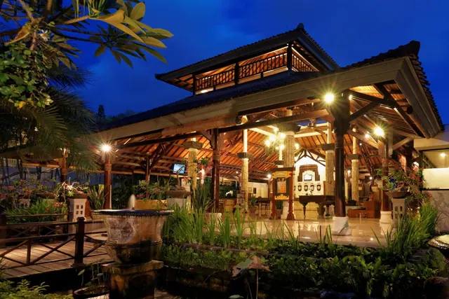 An illuminated traditional Balinese wooden pavilion with a tiled roof stands beside a planted pond at dusk.