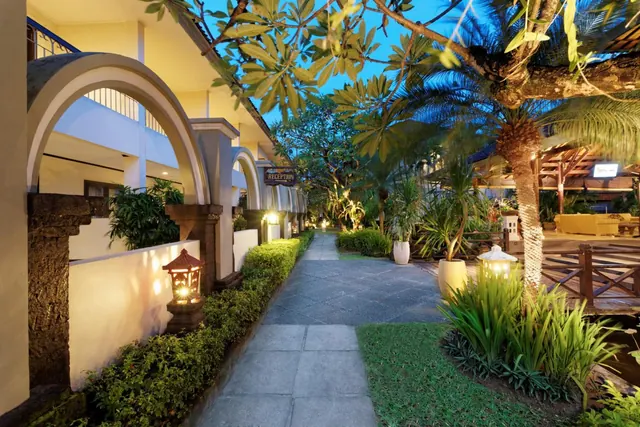 A lit garden pathway runs between arched hotel buildings with lanterns, shrubs, palm trees and a reception sign at dusk.