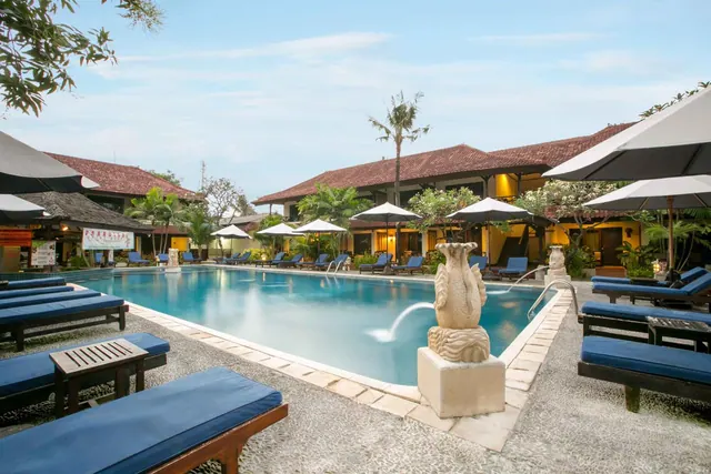 A rectangular swimming pool with blue sun loungers and white shade umbrellas in front of a two-storey building with a terracotta roof, with a stone sculpture at the pool edge.