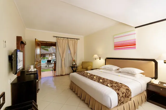 A bedroom with a king bed dressed with a brown runner and frangipani flowers, a wall-mounted television, an armchair, and an open door onto a pool outside.