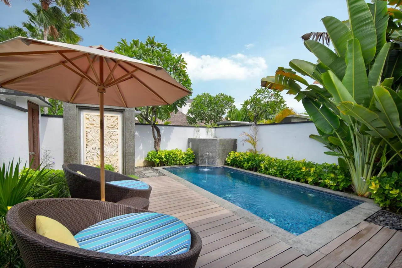 A small private pool with a wooden deck, wicker lounge chairs under a beige parasol, and banana plants along the white walls.
