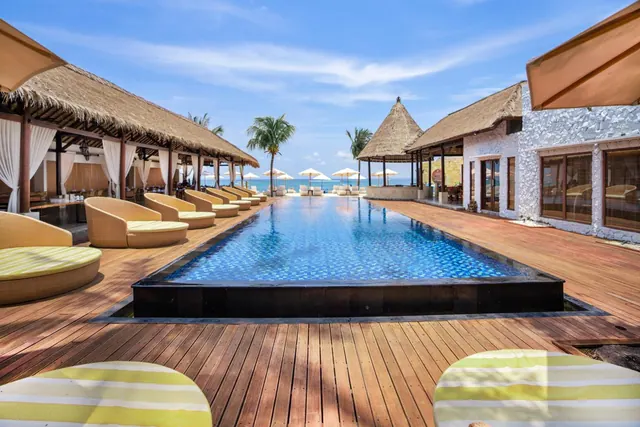A long rectangular swimming pool runs toward the sea, with thatched-roof cabanas, sun loungers and parasols on the wooden deck.