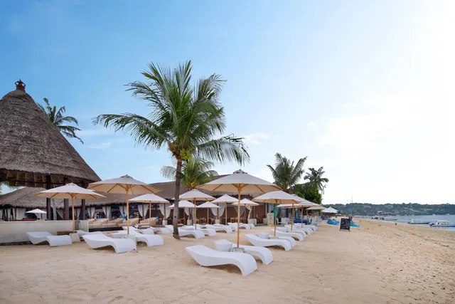 White sun loungers and cream parasols stand on a sandy beach beside palm trees and a thatched pavilion, with the sea and boats visible in the distance.