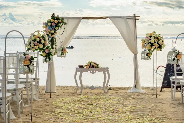 A beach wedding setup with a draped arch, flower arrangements, white chairs and rose petals on the sand, with the sea behind.