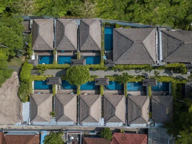 Aerial view of a row of thatched-roof villas, each with a small private pool, set among green trees.